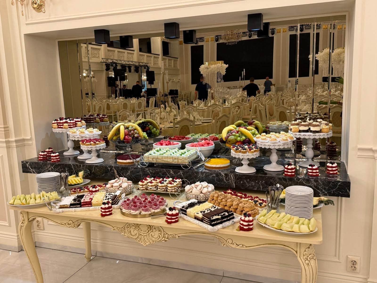 Masă de deserturi la un eveniment, cu tarte, macarons, pahare cu prăjituri și fructe proaspete aranjate pe stative de diferite înălțimi, într-o sală cu pereți oglindă