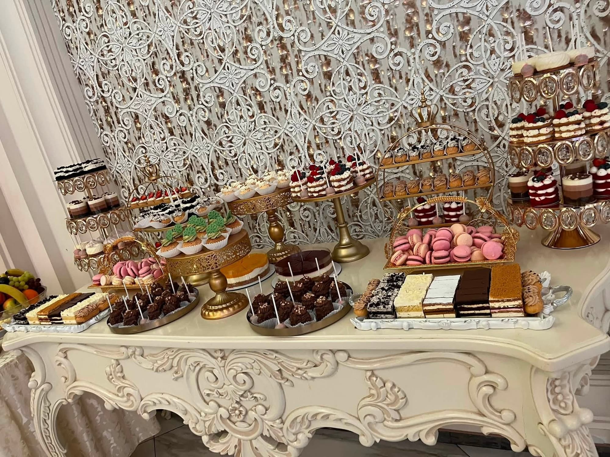 Masă de deserturi pe o consolă albă sculptată, cu felii de tort, macarons roz, prăjituri glazurate și platouri asortate, pe fundal cu perdea decorativă albă