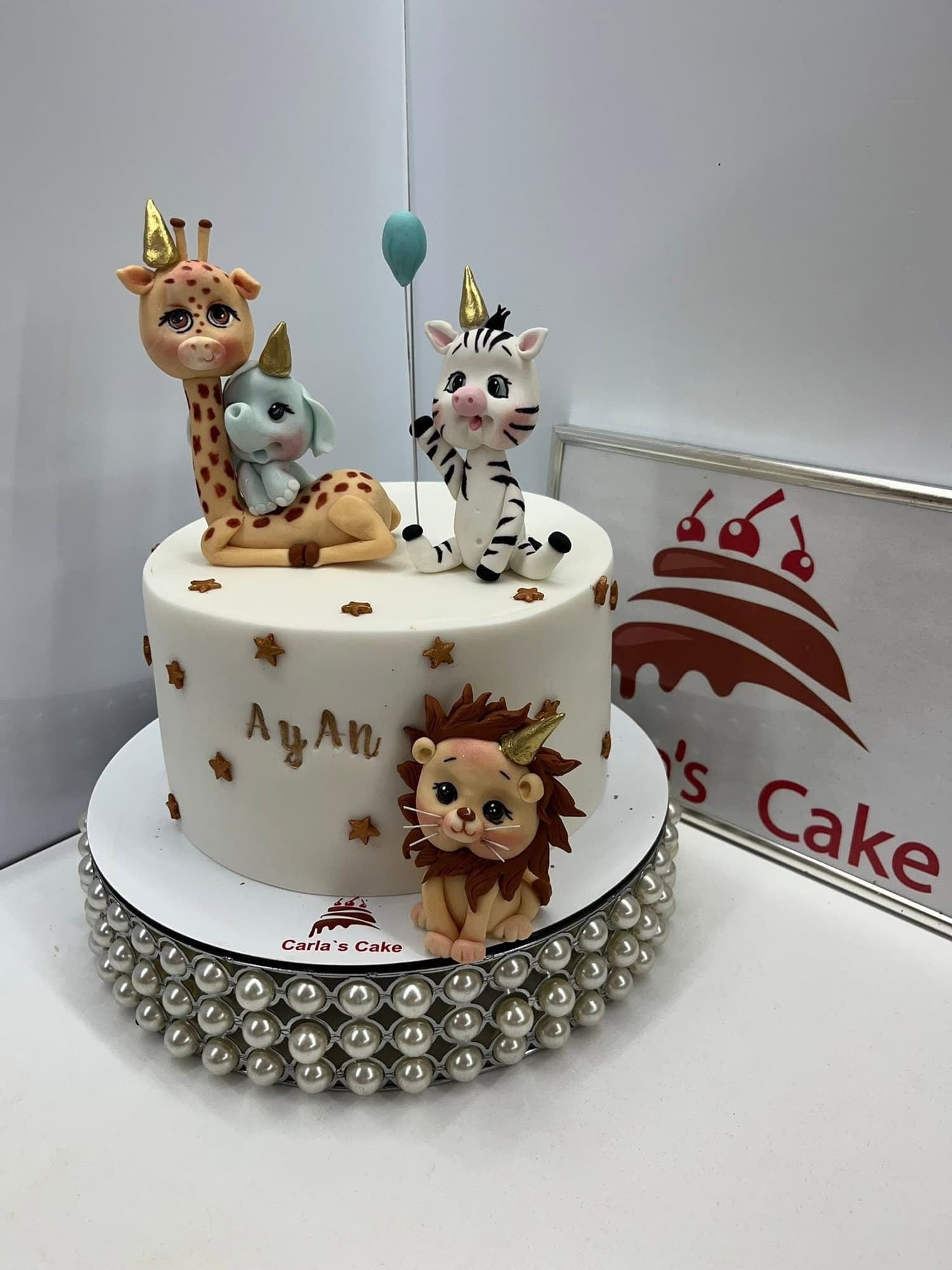 Tort aniversar pentru copii cu figurine fondant de animale safari — girafă, elefant, zebră și leu — și numele "AYAN" scris cu glazură aurie, pe suport cu perle argintii; siglă Carla's Cake vizibilă în fundal