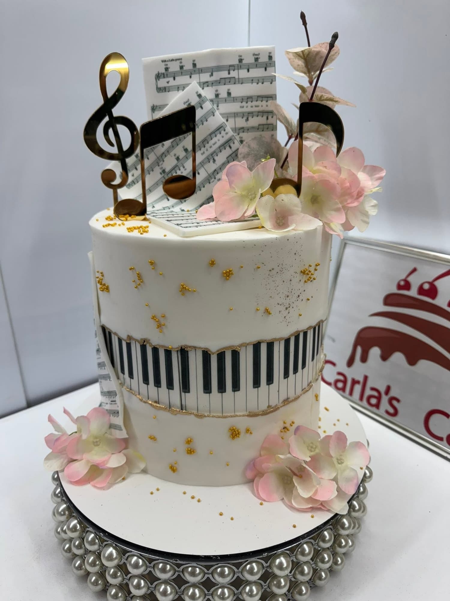 Tort aniversar cu temă muzicală — bandă fondant cu clape de pian, decor cu note muzicale pe partitură, topper auriu în formă de cheie sol și flori roz — pe suport cu perle argintii; siglă Carla's Cake vizibilă în fundal