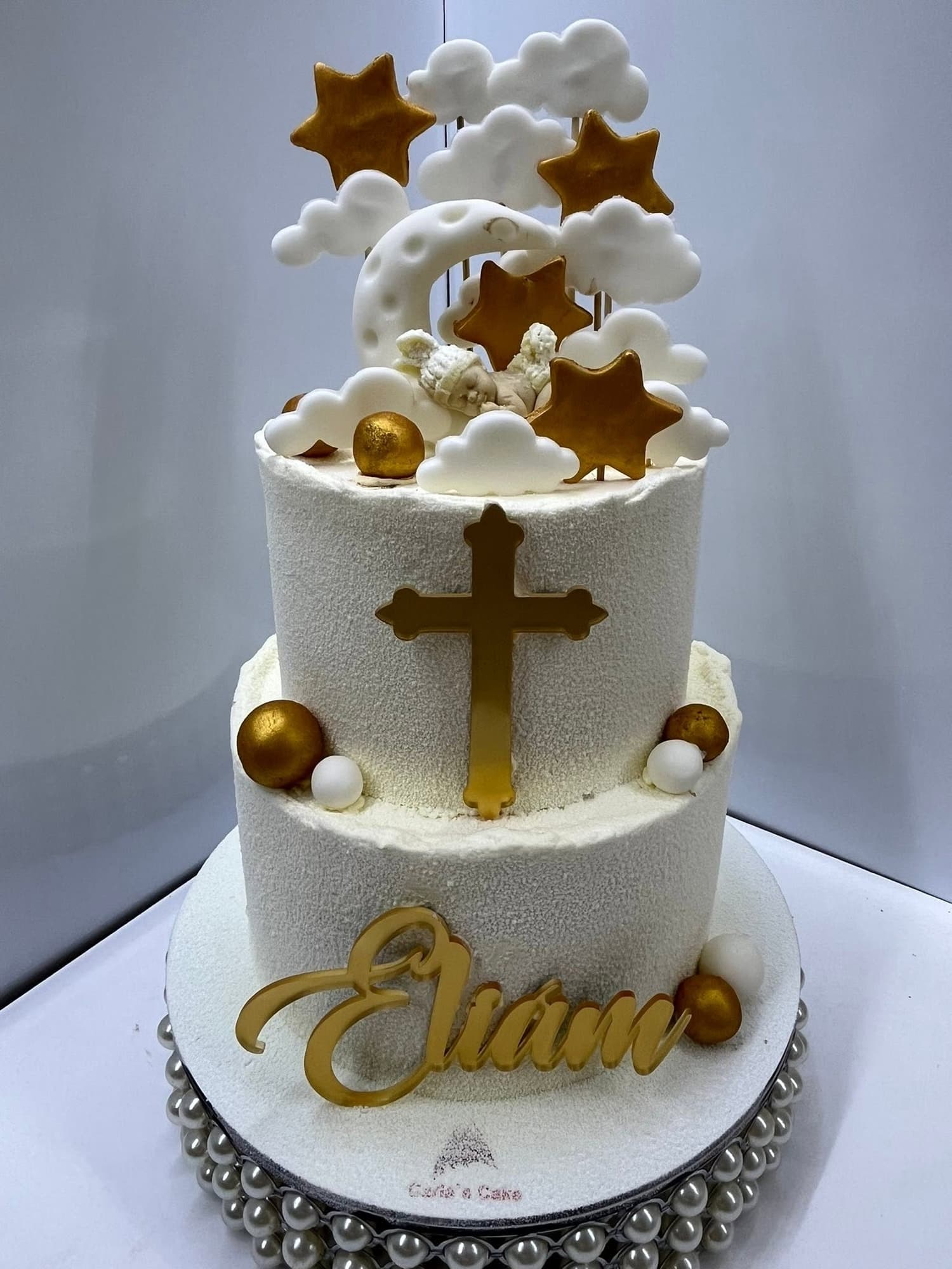 Tort de botez cu două niveluri, glazură albă texturată, o cruce aurie, stele și nori din fondant alb și numele copilului scris cu glazură aurie, pe suport cu perle argintii; siglă Carla's Cake pe platou