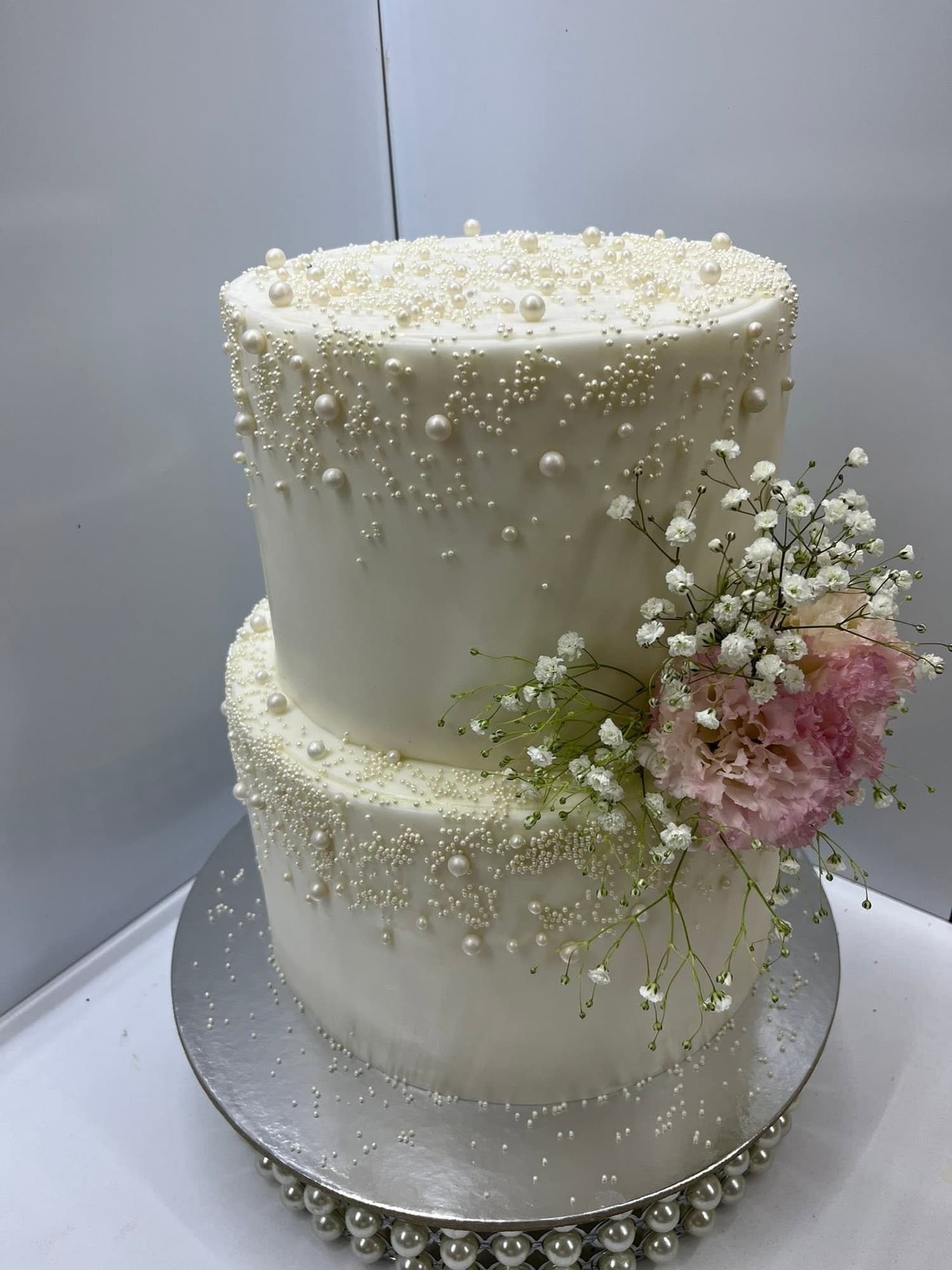 Tort de nuntă cu două niveluri, glazură albă decorată cu perle comestibile și un buchet de flori roz și albe proaspete, pe fundal gri de studio